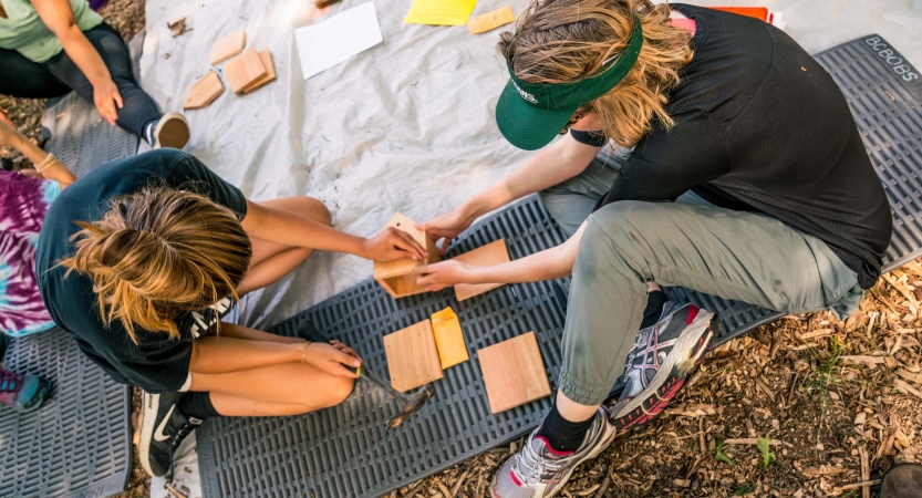 Two people sit on a pad on the ground and hold small pieces of wood during a service project. 